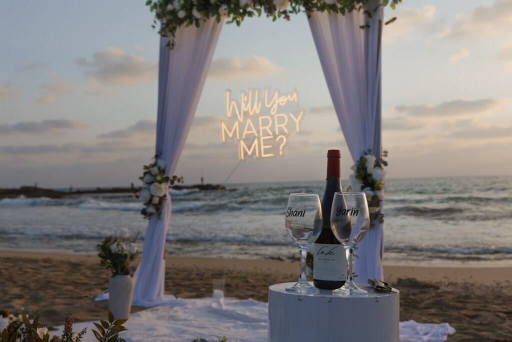 Néon LED avec la phrase Will You Marry Me sous un arche de mariage en bord de mer au coucher du soleil